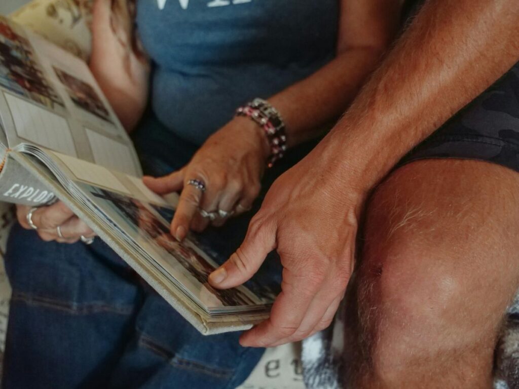 Close-up of a couple sitting together and looking through a photo album, pointing at pictures and holding the book on their laps.