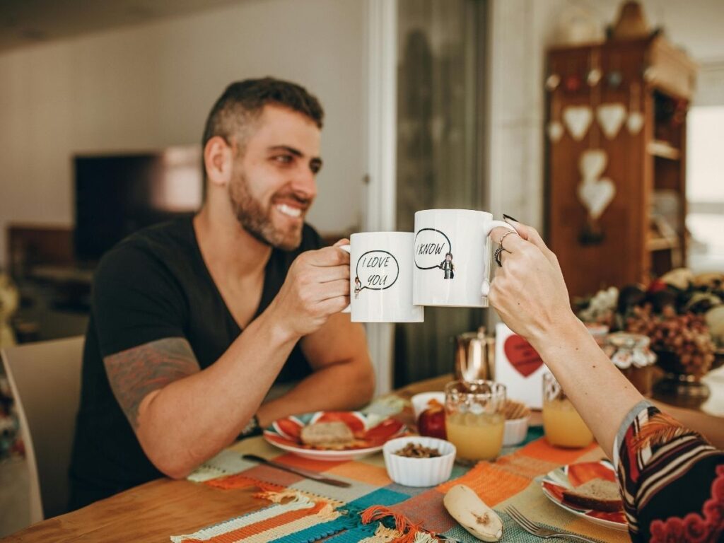 A couple sitting at a breakfast table, smiling and clinking matching mugs that read “I love you” and “I know,” enjoying a cozy meal together.
