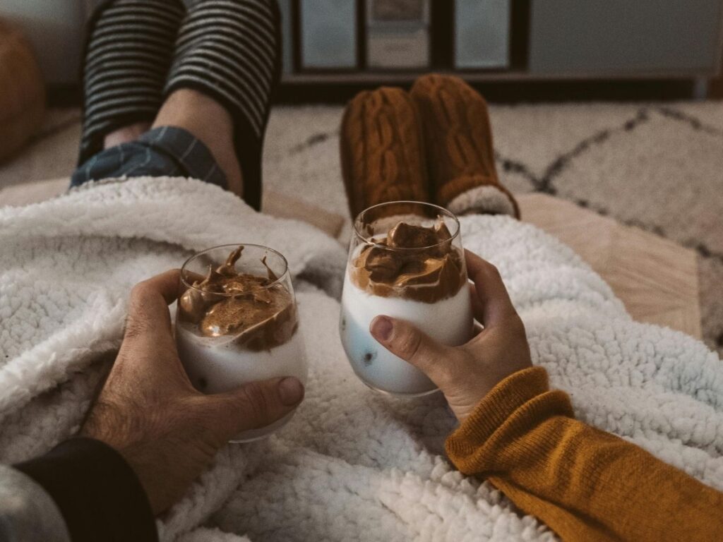 Two people relaxing under a blanket, holding chocolate desserts while watching a movie on TV in a cozy, candlelit living room.