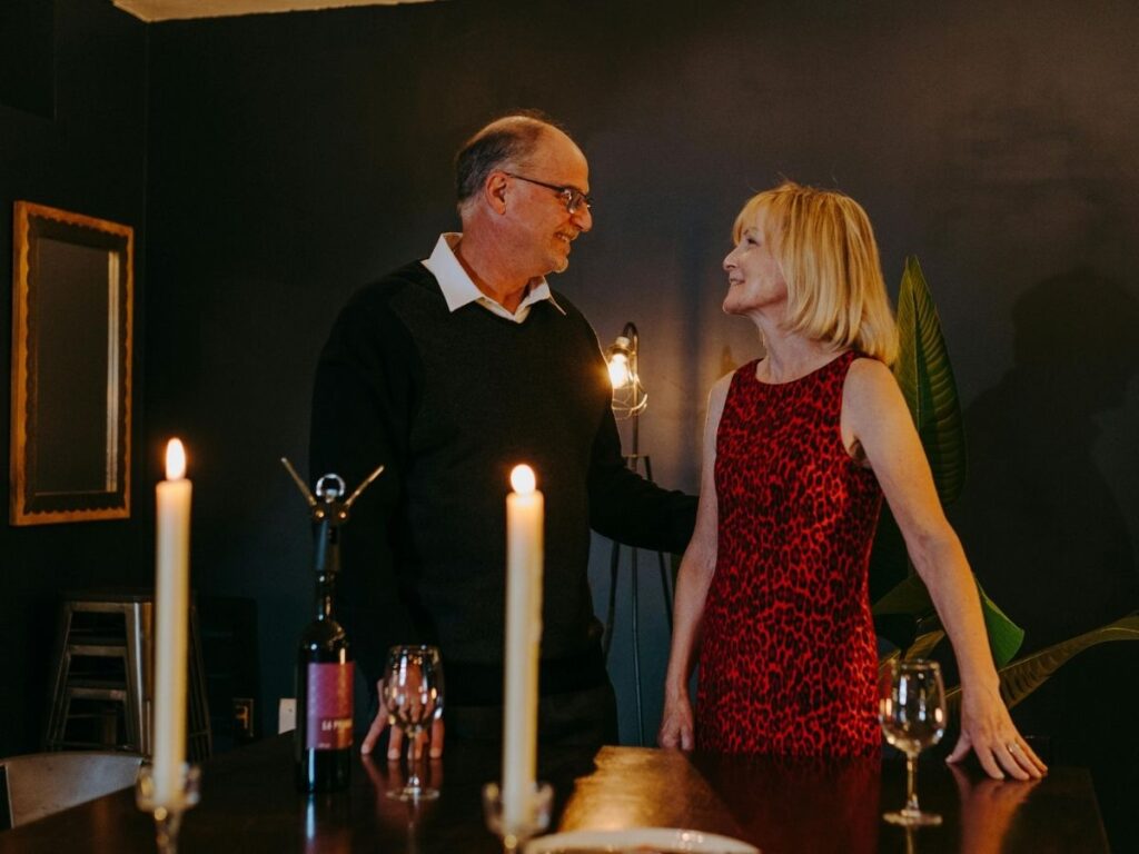 Couple standing by a candlelit table with wine glasses, smiling at each other.