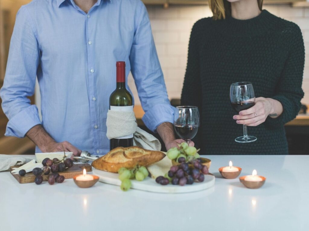 Couple holding wine glasses beside bread, grapes, and candles on a kitchen counter.