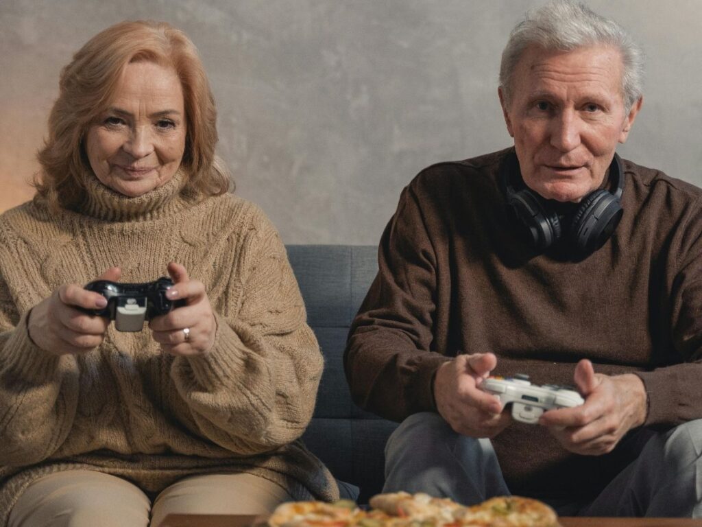 An older couple sitting side by side on a couch, each holding a game controller and looking focused while playing a video game. There’s a plate of food in front of them, and the man has headphones resting.