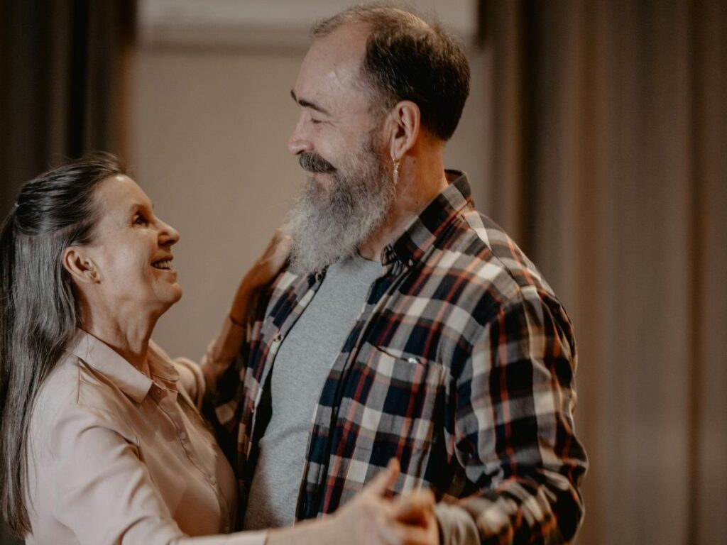 A middle-aged couple smiling at each other while slow dancing in a cozy living room.