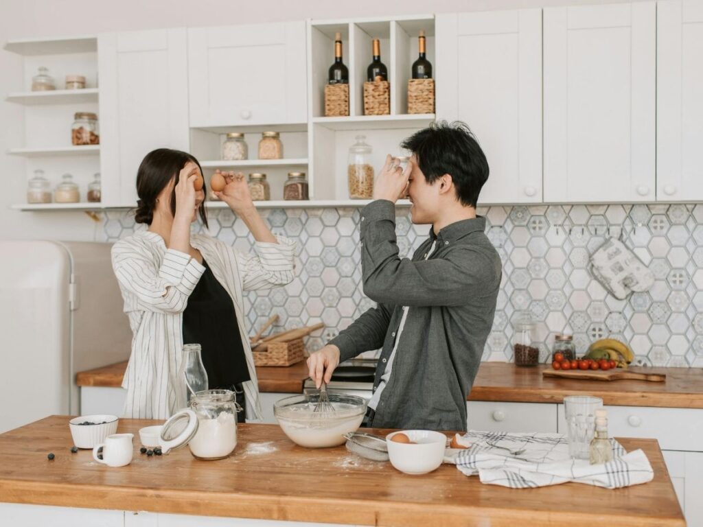 A couple playfully holding eggs to their foreheads while cooking together in a bright, modern kitchen.