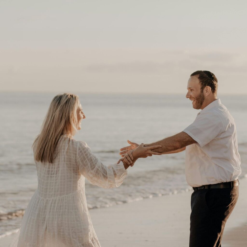 Couple holding hands and smiling while walking along the beach at sunset.