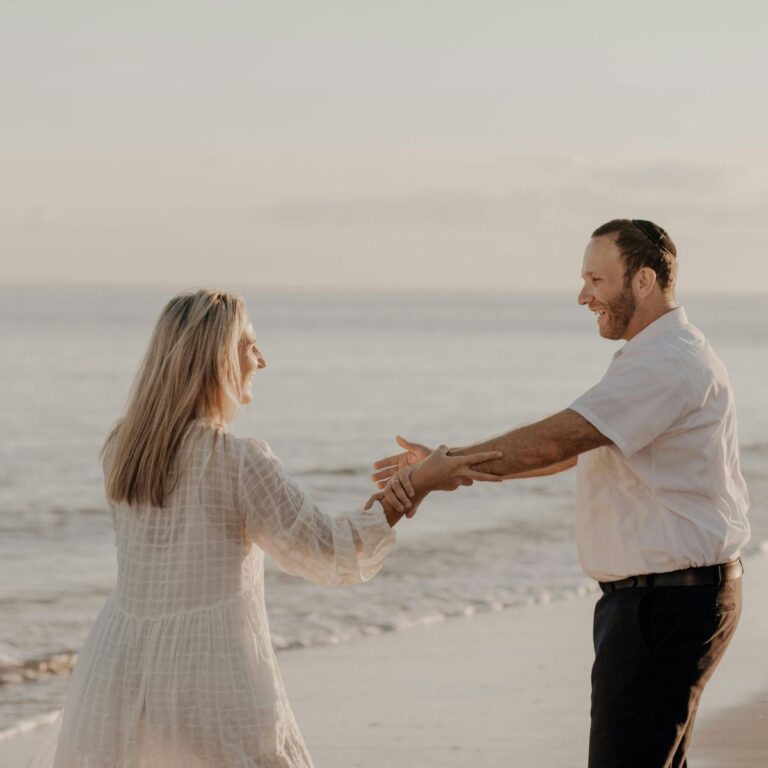 Couple holding hands and smiling while walking along the beach at sunset.