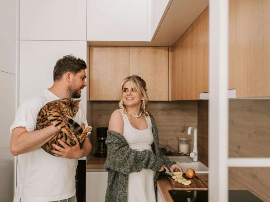 A couple standing together in a modern kitchen. The man is holding a spotted cat, while the woman is smiling at him as she slices apples on a cutting board.