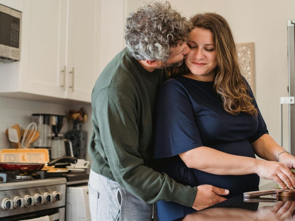 A couple standing together in a kitchen. The man gently kisses the woman on the cheek from behind while she prepares food at the counter.