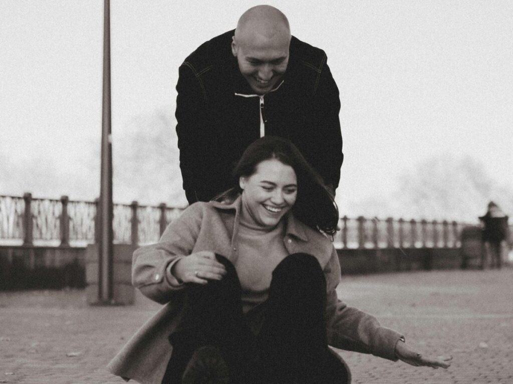 A man pushing a smiling woman riding a skateboard on a paved walkway.