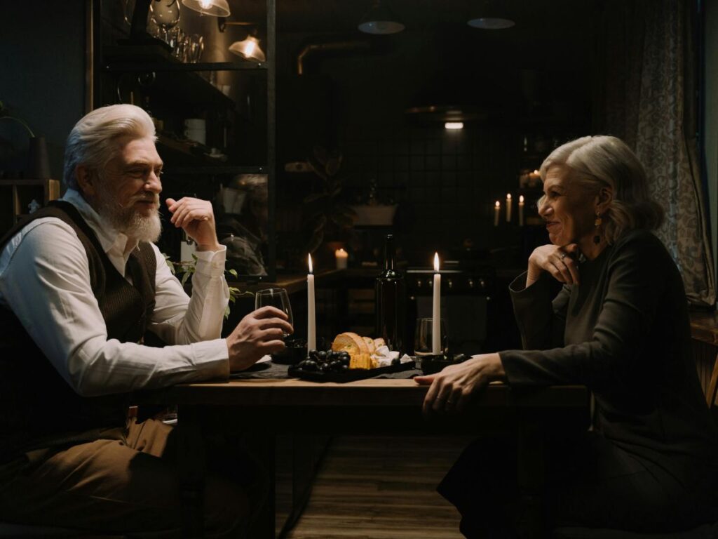 An older couple sharing a candlelit dinner at home, sitting across from each other with wine, bread, and soft warm lighting creating an intimate evening atmosphere.