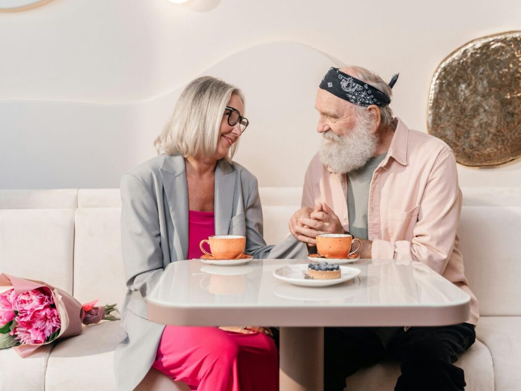 An older couple sitting across from each other at a small café table, holding hands and smiling warmly. There are two cups of coffee and a small dessert on the table, and a bouquet of pink flowers rests beside them on the seat.