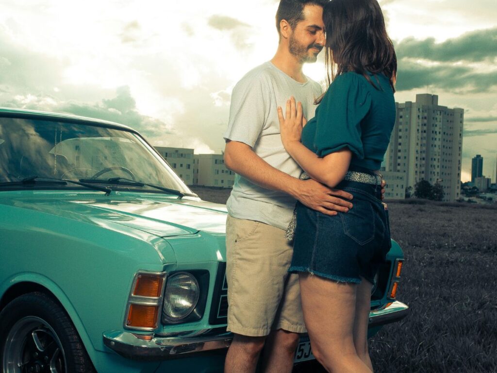 Couple embracing beside a vintage turquoise car at sunset with city buildings in the background.
