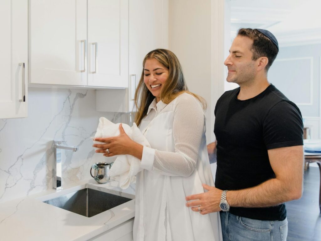 Smiling couple standing by a kitchen sink, with the man gently holding the woman as she dries dishes.