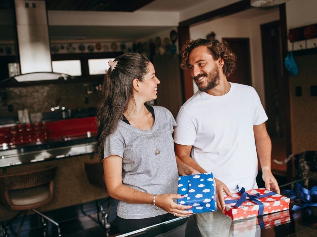 A smiling couple standing in their kitchen, looking at each other while holding wrapped gift boxes on a table.