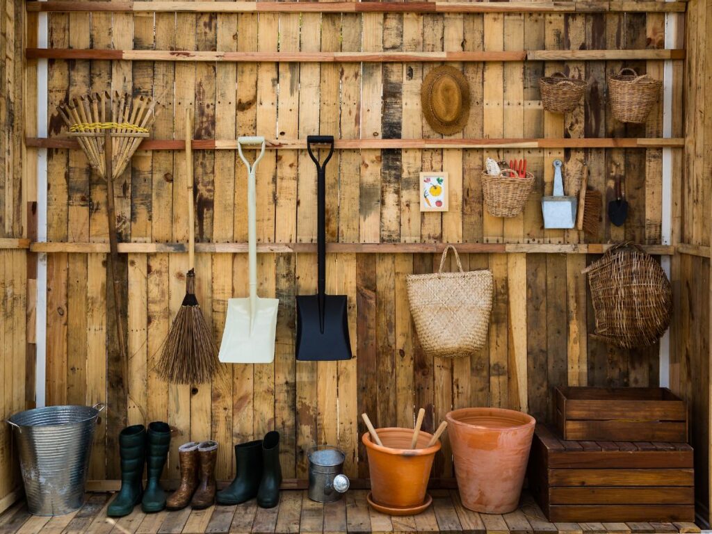 Garden tools and baskets neatly hung on a wooden shed wall, with boots, pots, and watering can arranged below.