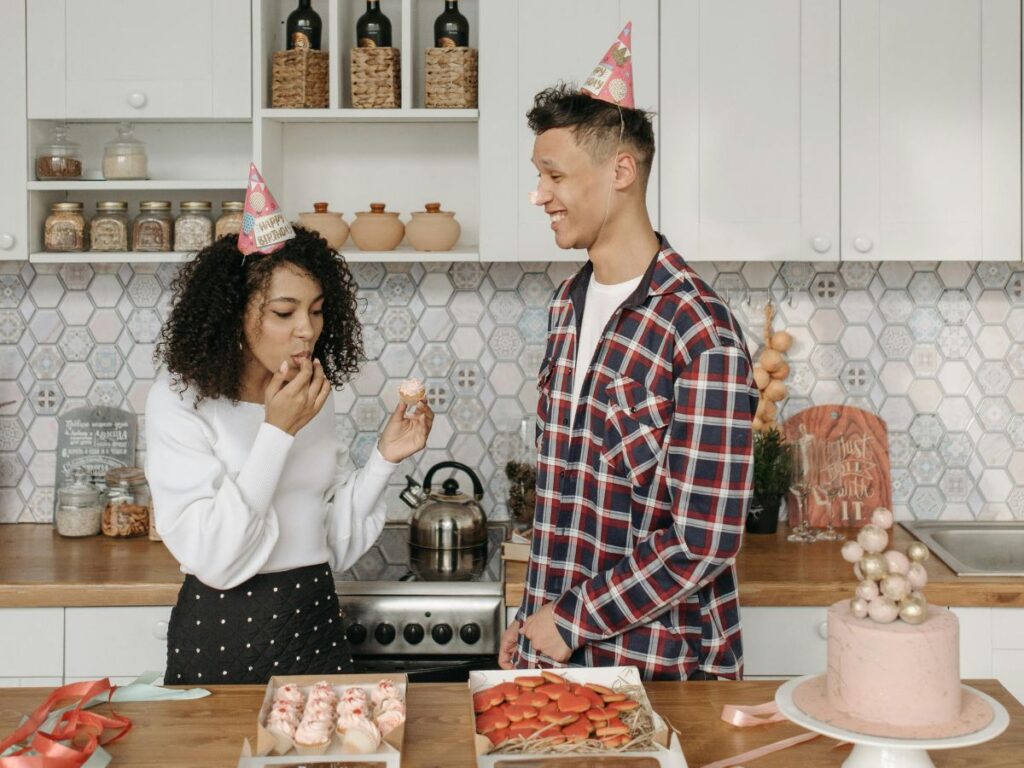 A couple wearing party hats tasting cupcakes together in a bright kitchen decorated for a birthday celebration.