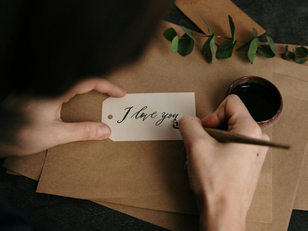 A person writing “I love you” in elegant script on a small tag, with envelopes, greenery, and an ink pot nearby.