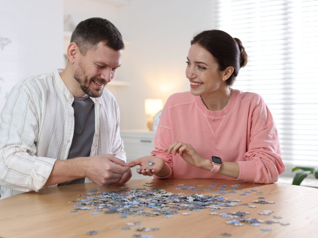 A smiling couple sitting at a table, working together on a jigsaw puzzle and sharing pieces as they chat.