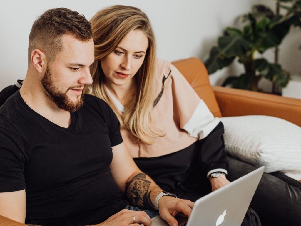 A couple sitting on a couch together, looking at a laptop screen and browsing online.