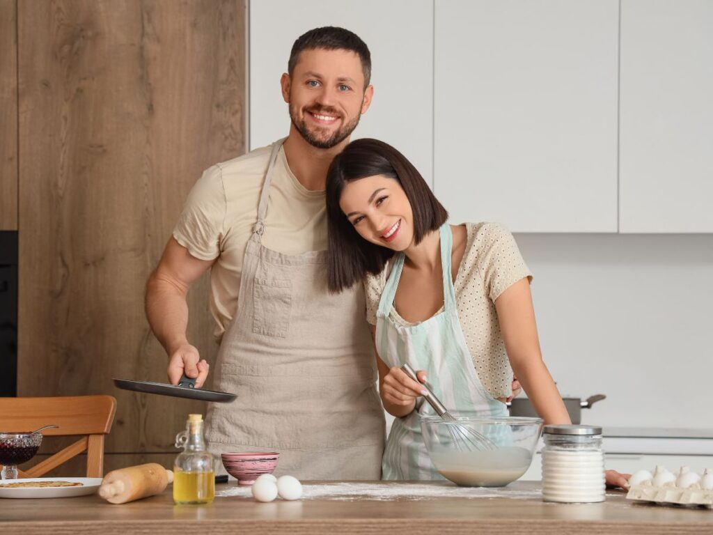 A smiling couple in aprons cooking together in a bright kitchen.