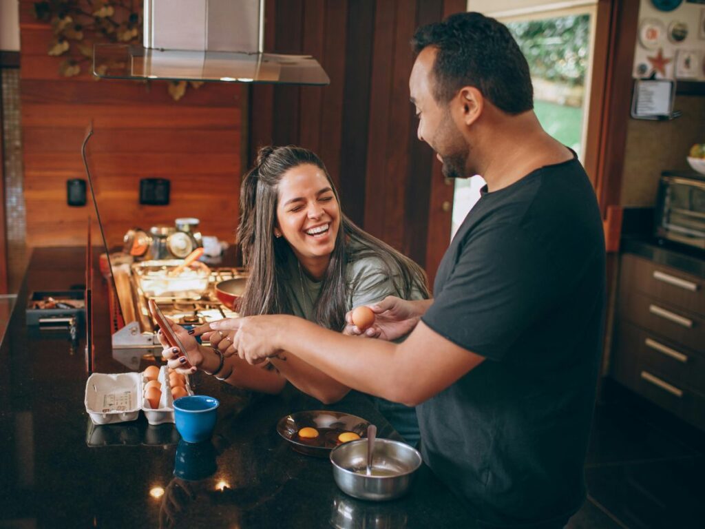 A couple laughing together in the kitchen while cracking eggs and cooking at the stove.