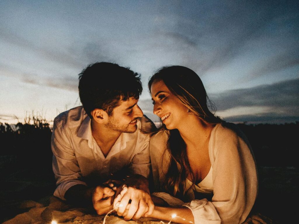 Couple lying close together outdoors at dusk, smiling at each other with soft string lights glowing around them under the evening sky.