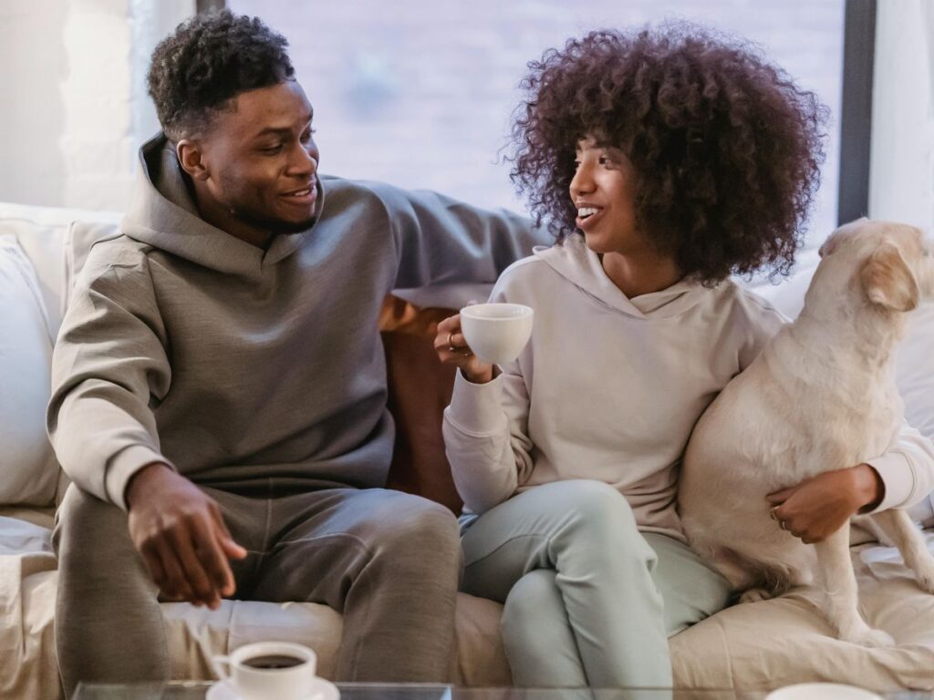 Couple sitting on a couch with their dog, smiling at each other while holding coffee in a bright living room.