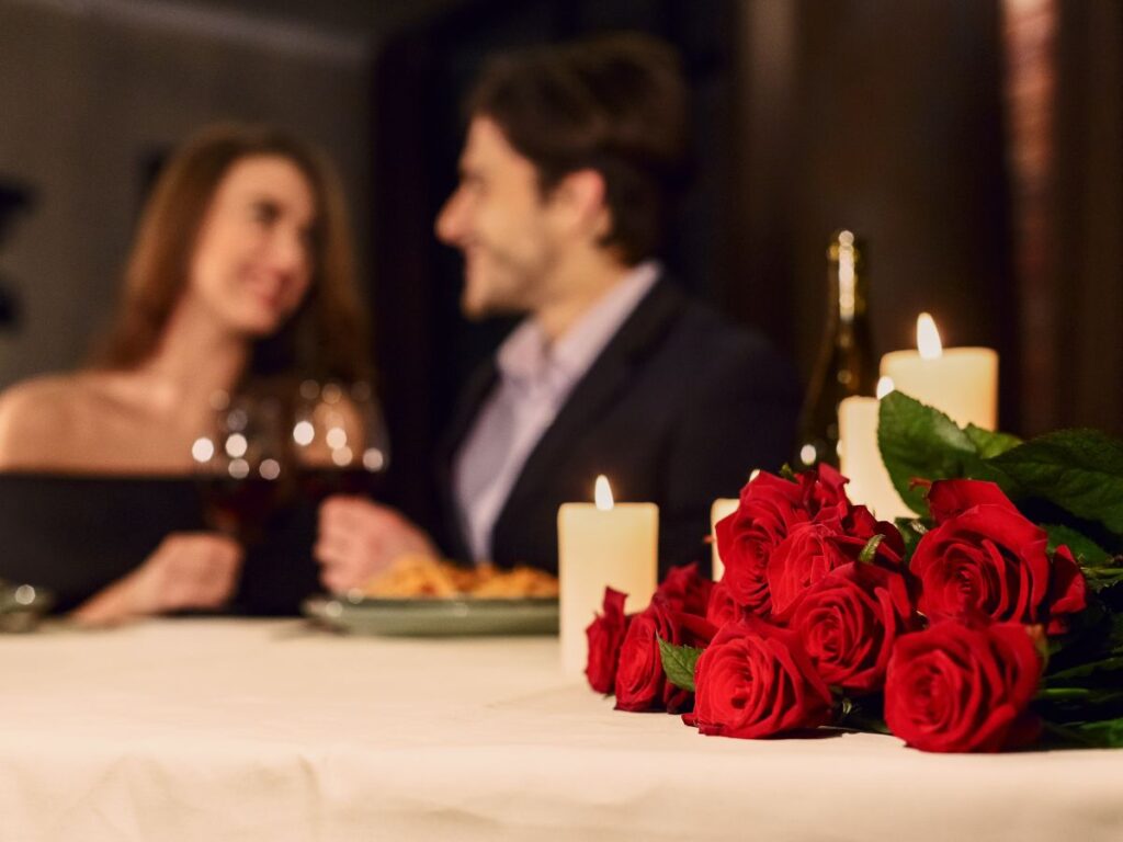 A couple enjoying a romantic candlelit dinner, holding wine glasses with red roses and candles on the table in the foreground.