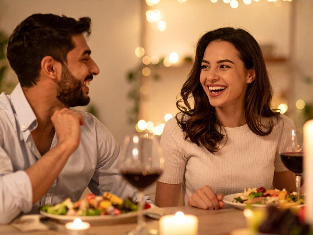 A smiling couple enjoying a cozy candlelit dinner at home, laughing together over plates of food and glasses of wine with warm string lights in the background.