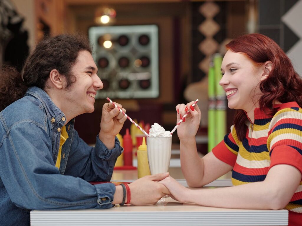 A smiling couple sitting across from each other at a diner, sharing a milkshake with two straws and holding hands.