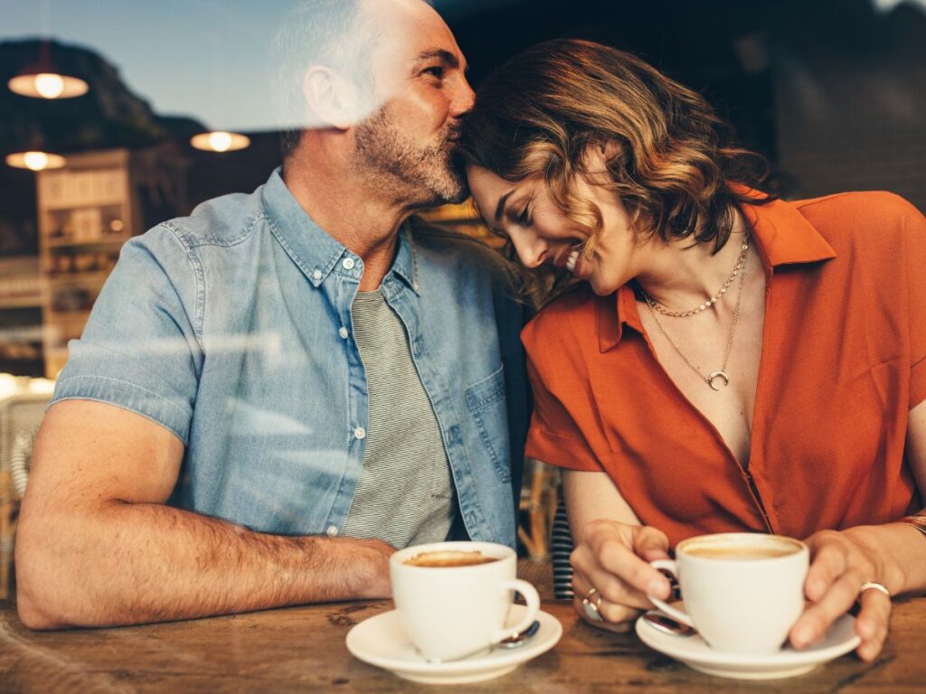 A cozy couple sitting side by side at a café table, sharing a quiet moment over two cups of coffee, with the man gently kissing the woman’s forehead as she smiles and leans into him.