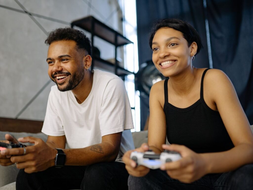 A smiling couple sitting on a couch playing video games together, each holding a controller and leaning forward with excitement.
