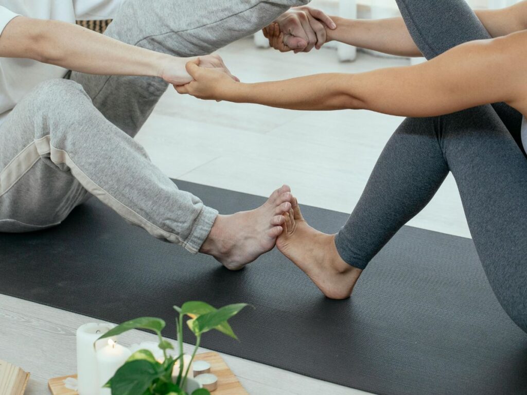 Two people sitting on yoga mats facing each other, holding hands and pressing their feet together in a partner stretch exercise, with candles and a small plant nearby.