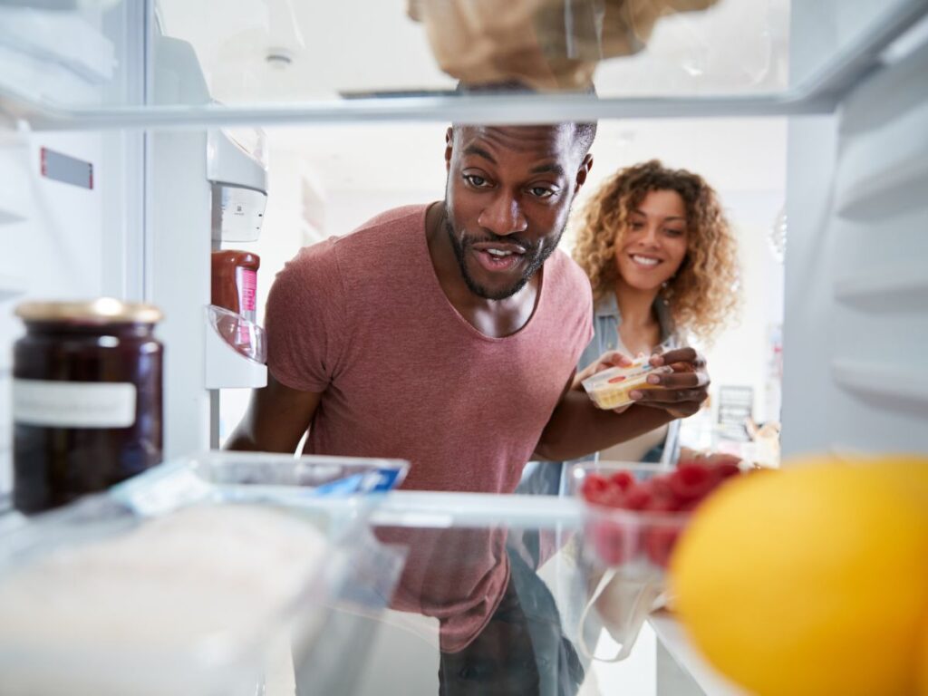 View from inside a refrigerator of a smiling couple looking in, with fruit and containers on the shelves in the foreground.