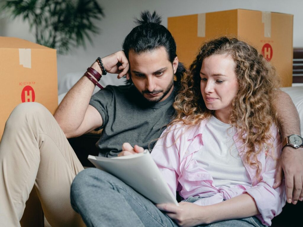 A couple sitting on the floor among moving boxes, reviewing papers together. The woman holds a clipboard while the man leans in with his arm around her, both focused and thoughtful, as if planning or organizing something in their new space.
