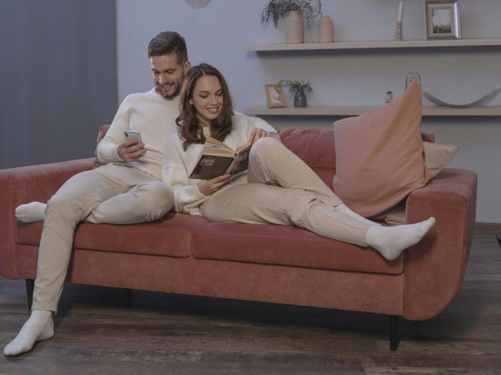Couple relaxing on a sofa at home, the woman reading a book while the man holds a phone and smiles beside her.