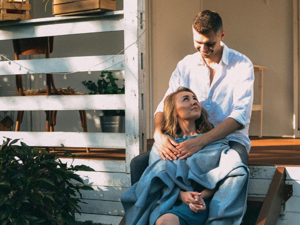 A couple sitting on wooden porch steps, wrapped in a blanket, sharing a quiet and affectionate moment outdoors.