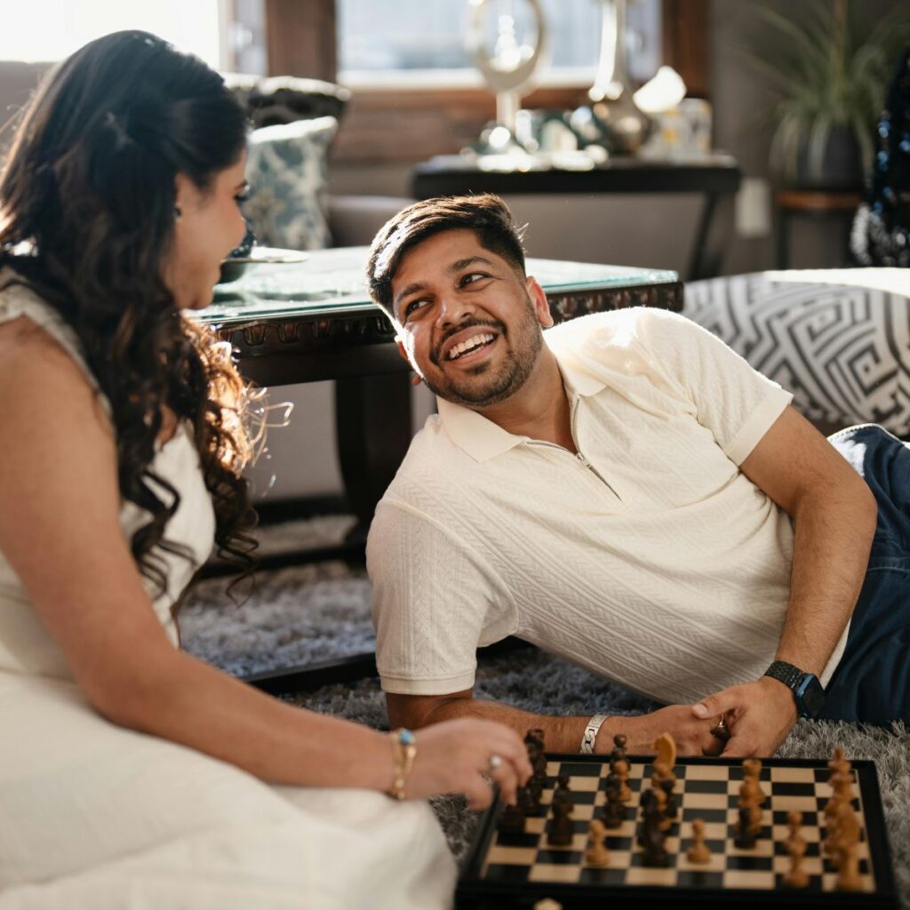 A couple relaxing on the floor at home, smiling at each other while playing a game of chess.