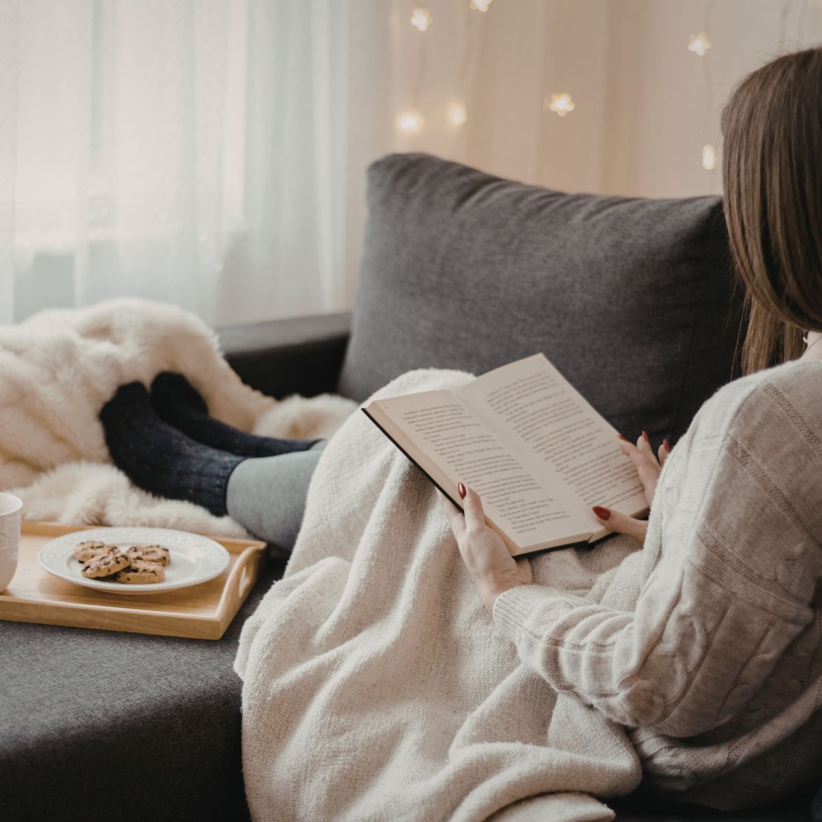 A cozy scene of a woman relaxing on a couch under a blanket, reading a book. A tray with cookies and a cup sits nearby, and soft string lights glow in the background.