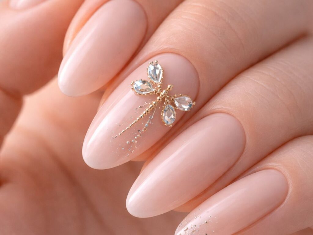 Close-up of almond-shaped soft nude nails with a glossy finish, featuring a detailed gold and crystal dragonfly accent on one nail, with subtle shimmer details against a clean, neutral background.