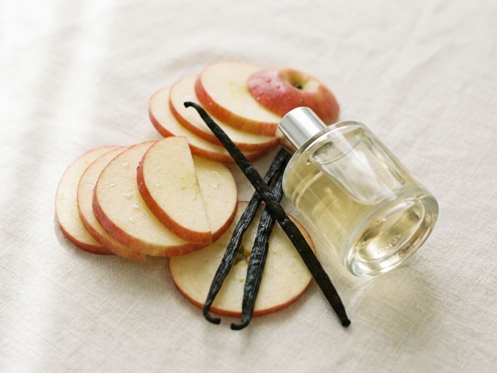 Perfume bottle with sliced apples and vanilla pods on neutral fabric