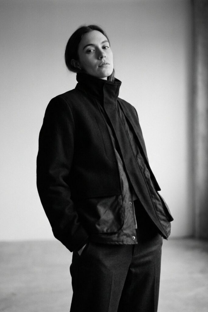 Black-and-white portrait of person in layered dark jacket standing by window