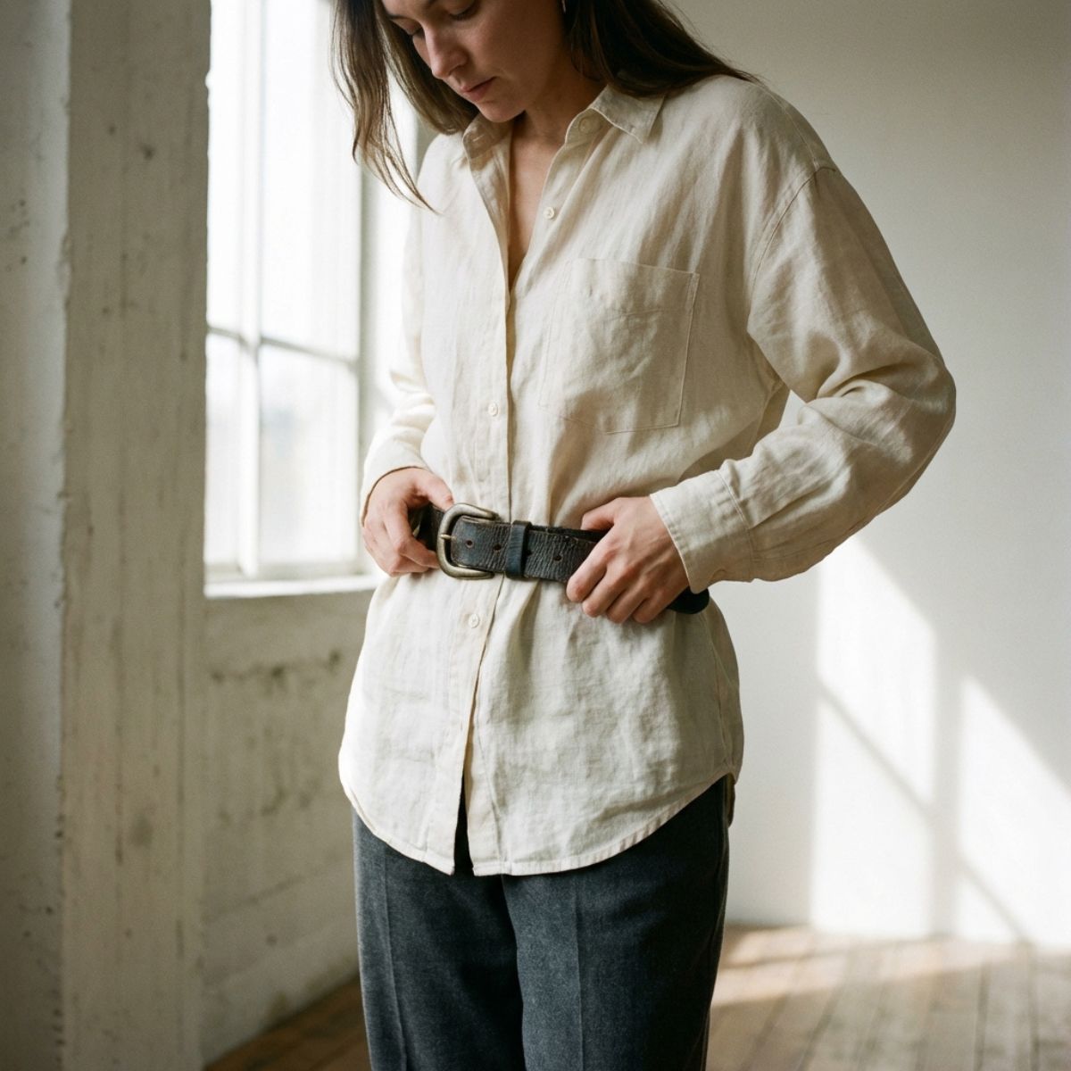 Close-up of a person styling a chunky belt over a loose beige button-down shirt in soft natural window light