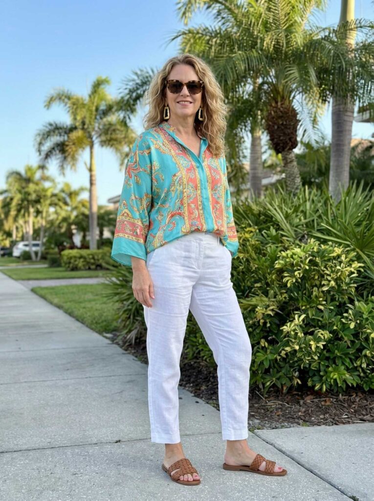 A woman in her 50s wearing a colorful printed blouse with white ankle-length pants and sandals, standing on a sunny Florida sidewalk with palm trees in the background.