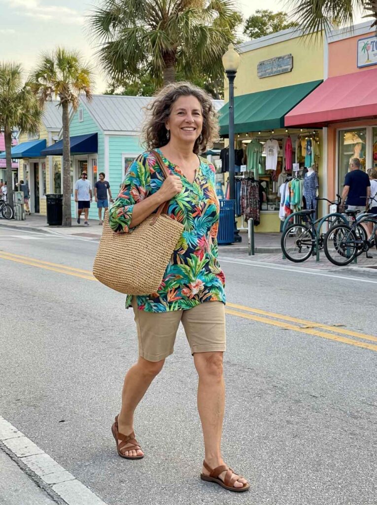 A woman in her 50s walking through a Florida street wearing a tropical print top, knee-length shorts, sandals, and carrying a woven tote bag.