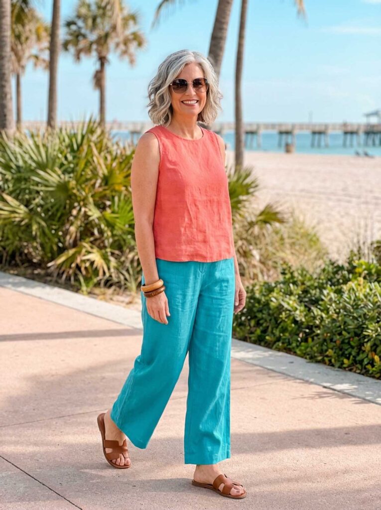 A woman in her 50s wearing a sleeveless coral top with wide-leg teal pants and sandals, walking near a sunny Florida beach path.