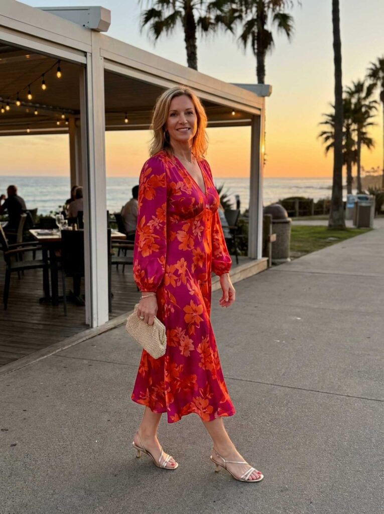 A woman in her 50s wearing a bold floral midi dress and heels walking near a coastal restaurant at sunset in Florida.
