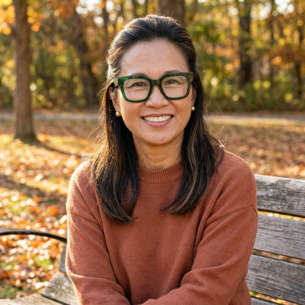 Asian woman in her 50s with dark highlighted hair half up half down and bold forest green glasses sitting on a park bench in autumn.