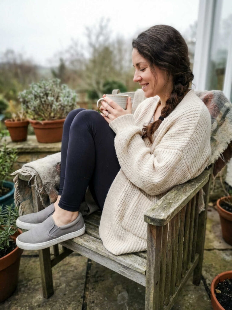 Woman in an oversized cardigan and leggings curled up on a patio chair with coffee.