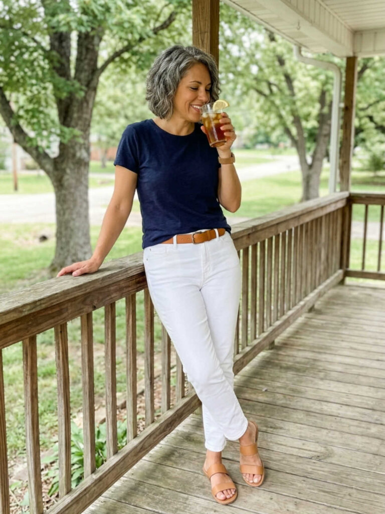 Woman in a navy tee and white jeans sipping iced tea on a porch railing.
